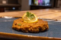 A single breaded, fried Tuna Cake on a dark blue plate, garnished with a lemon slice and parsley.