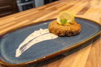 Golden breaded Tuna Cake topped with a lemon slice and herb sprig on a blue plate with sauce swipe.