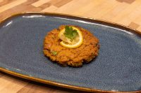 A golden-brown Tuna Cake topped with a lemon slice and herb sprig on a dark blue oval plate