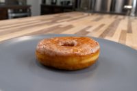 Donuts and Donut Holes A glazed ring donut from Donuts and Donut Holes sits on a dark gray plate on a wooden counter.