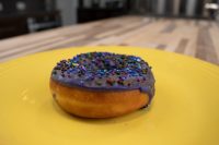 A single purple-glazed donut with rainbow sprinkles from Donuts and Donut Holes, on a yellow plate.
