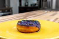 Donuts and Donut Holes A single Donuts and Donut Holes pastry with purple glaze and rainbow sprinkles on a yellow plate.
