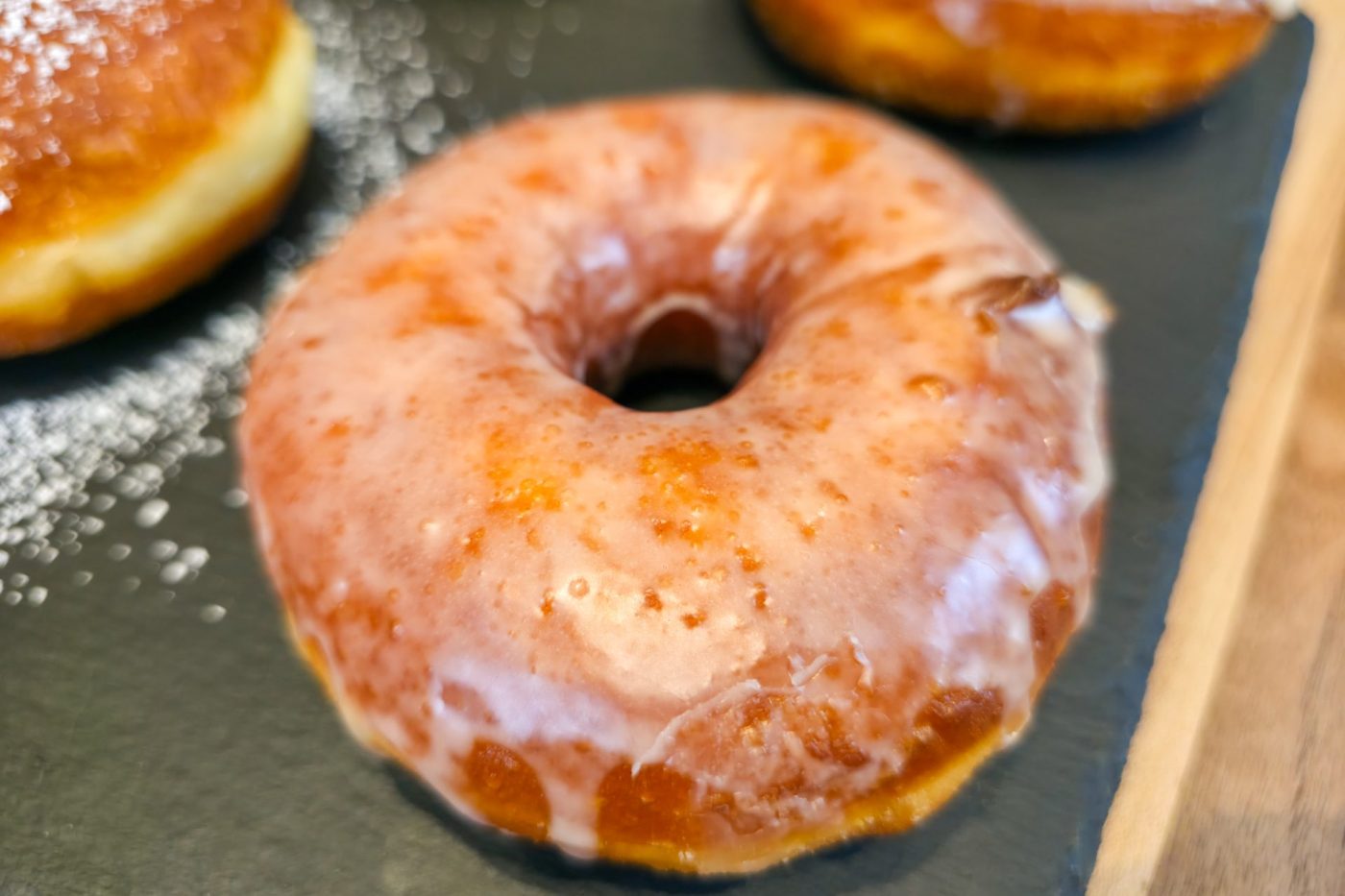 Close-up of a glazed doughnut from Donuts and Donut Holes on a dark slate surface, with two more doughnuts partly visible.