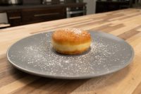 Czech Filled Donuts Koblihy A golden Czech Donuts Koblihy dusted with powdered sugar on a gray plate atop a wooden countertop.