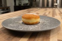 Czech Filled Donuts Koblihy A fried Czech Filled Donuts Koblihy dusted with powdered sugar, filling visible at its seam, on a grey plate.