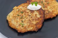Potato Pancakes Bramboraky Two crispy Potato Pancakes Bramboraky on a dark plate, topped with sour cream and chopped scallion.