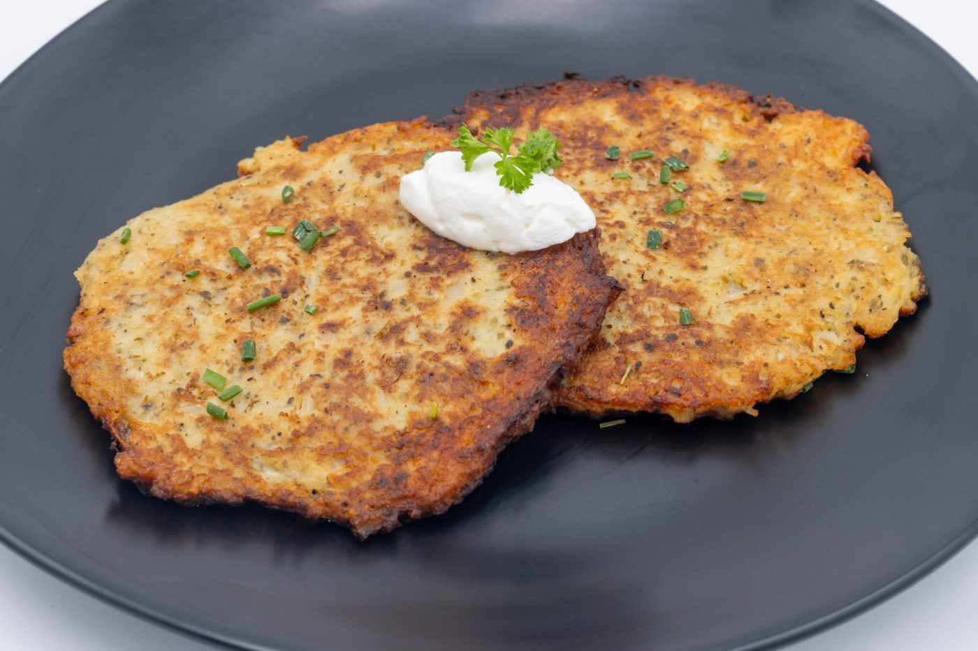 Potato Pancakes Bramboraky Two crispy Potato Pancakes Bramboraky on a black plate with sour cream and chopped chives on top.