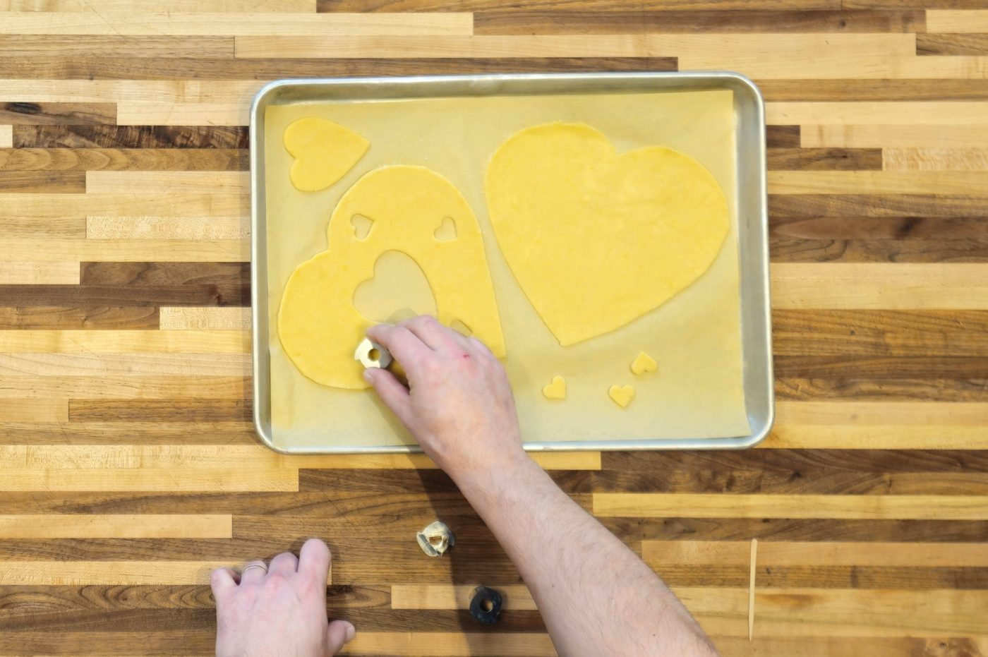Hand pressing a heart cutter into rolled dough for Linzer Tart Hearts on a lined baking sheet.