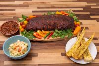 Glazed rack of American BBQ – Honey Pork Ribs on a board with peppers, herbs, coleslaw, beans, and garlic bread.