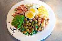 Cobb Salad Cobb Salad on a white plate with ham, egg halves, cheeses, avocado, olives, and croutons over greens.