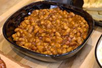Easy Baked Beans Dark square bowl filled with baked beans coated in thick brown sauce, with bread visible at the edge.
