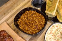 Easy Baked Beans Bowl of Baked Beans in a rich brown sauce, served on a wooden board with garlic bread and coleslaw nearby.