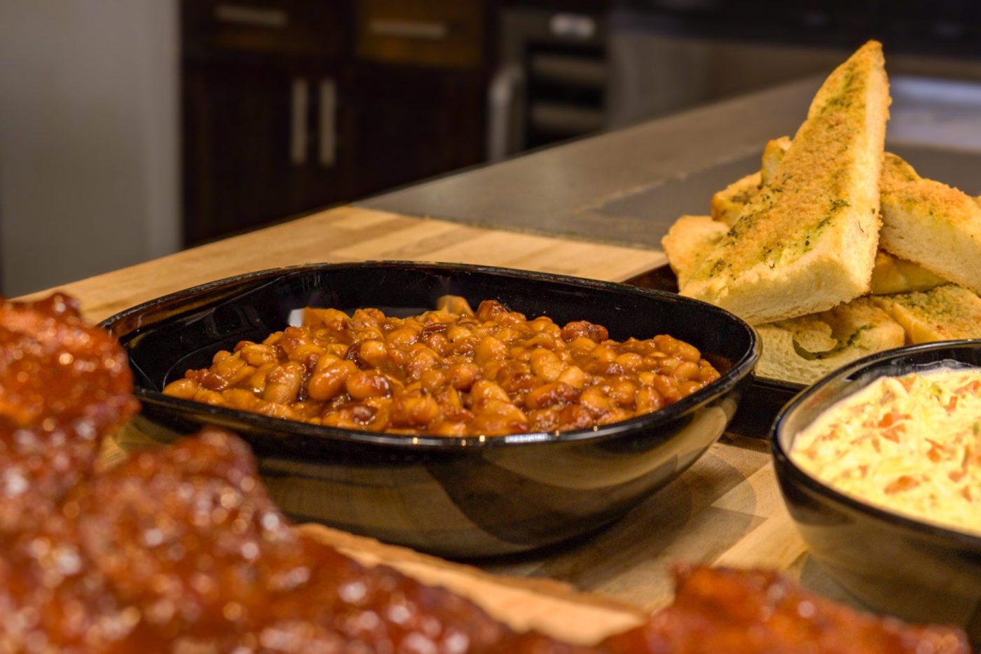 Easy Baked Beans Black dish of Baked Beans in reddish sauce, served with garlic bread, a creamy side, and ribs.