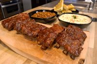American BBQ Honey Ribs Glazed American BBQ – Honey Pork Ribs on a wooden board, with baked beans, coleslaw, and bread slices behind