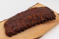 A charred rack of American BBQ – Honey Pork Ribs glazed dark brown, resting on a wooden board.