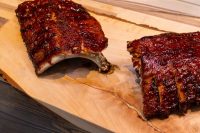 Two racks of American BBQ – Honey Pork Ribs with dark glaze, one sliced, on a wooden board.