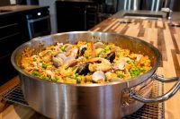 Large steel pan of Paella with saffron rice, peas, clams, mussels, and shrimp on a wooden counter.